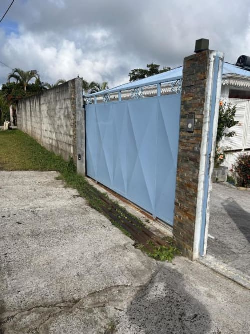 Portail en métal bleu d'accès à la Résidence B&L Lagon à Sainte-Luce, Martinique, avec piliers en brique et vue sur le jardin