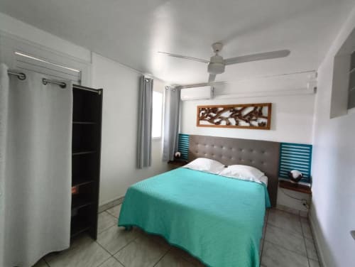 Chambre lumineuse à la Résidence B&L Lagon avec literie turquoise, tête de lit grise, ventilateur et décor moderne à Sainte-Luce