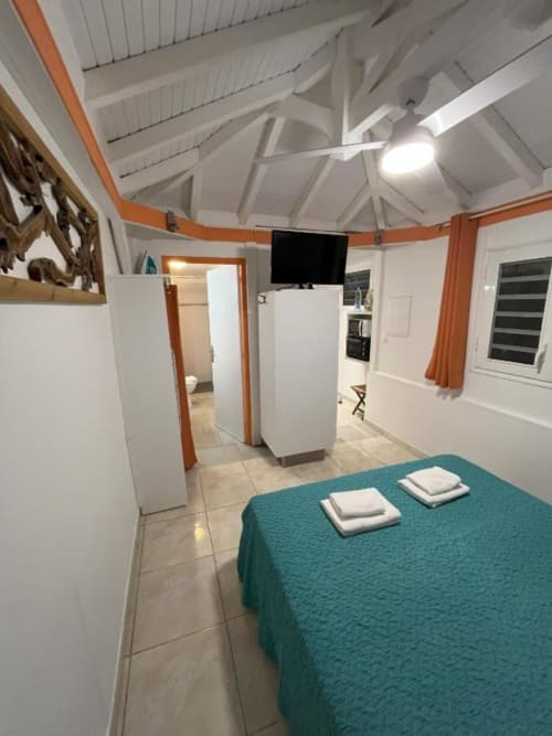 Chambre moderne à la Résidence B&L Lagon à Sainte-Luce avec lit turquoise, murs blancs, poutres orange, ventilateur et écran plat