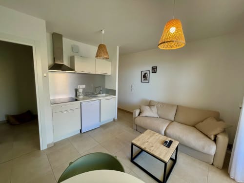 Modern open-plan studio at Résidence Marina di Bravone with equipped kitchenette, beige sofa, wooden table, and woven pendant