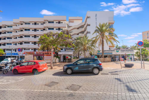 Straßenansicht des Apartmentkomplexes Home Stay Gran Canaria in Maspalomas mit Palmen, parkenden Autos und Supermarkt