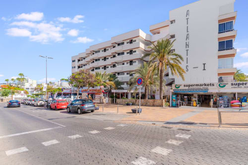 Atlantis-Apartmentgebäude in Maspalomas mit Palmen, Straßenparkplatz und Supermarkt im Erdgeschoss an sonniger Küstenlage