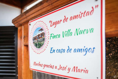 Cartel de bienvenida de Finca Villa Nueva en Mala, España, con la leyenda 'Lugar de amistad', vistas a la montaña y agradecimiento a José y