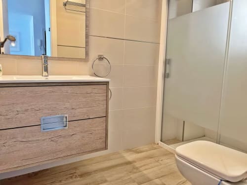 Baño moderno en apartamento frente al mar en Fuengirola con mueble de madera, azulejos blancos y ducha con mampara de cristal