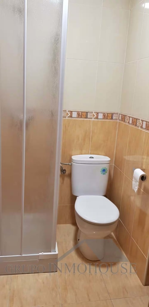 Baño moderno con inodoro blanco, azulejos crema y beige, cabina de ducha y papel higiénico en Grupo Inmo House Fuengirola