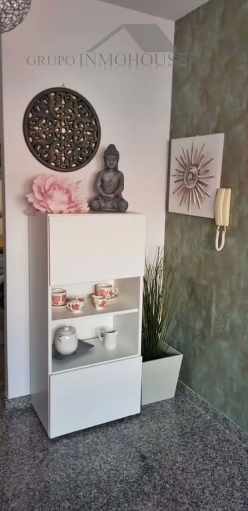 Entrada moderna de apartamento en Grupo Inmo House Fuengirola con estantes blancos, estatua Buda, flor rosa y verde salvia