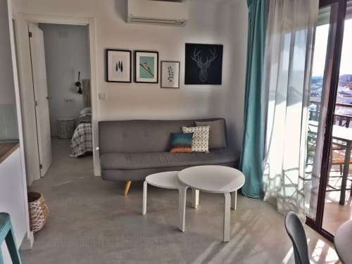 Apartamento moderno en Fuengirola con sofá gris, mesas blancas, arte mural y vistas al mar y ciudad desde el balcón