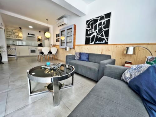 Moderno salón en Fuengirola con sofá gris, panelado de madera, área de cocina y arte contemporáneo.