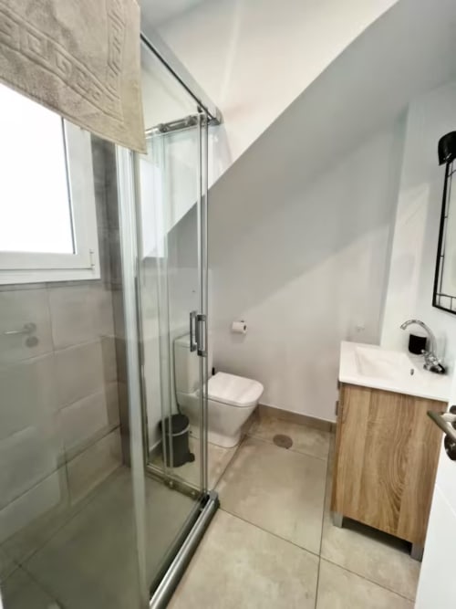 Baño moderno en apartamento de Fuengirola con ducha de cristal, inodoro blanco, tocador de madera y techo inclinado