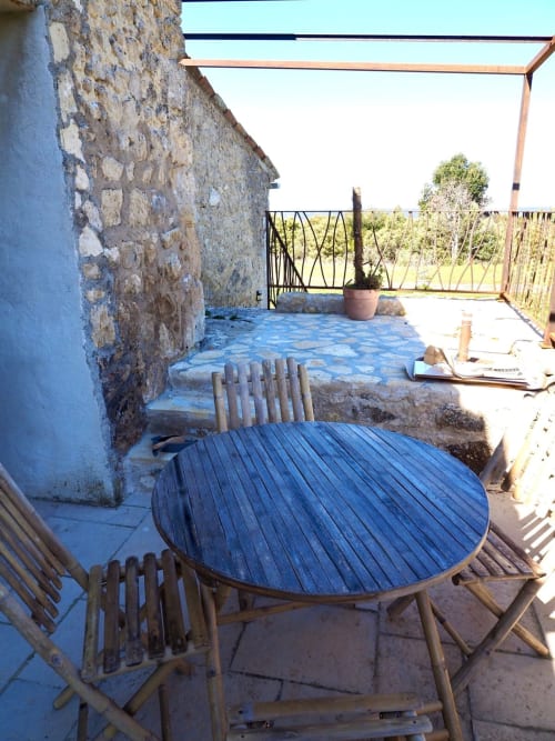 Terrasse rustique de la Bastide Saint Michel avec table ronde en bois, chaises, murs en pierre et vue sur le jardin à Baudinard sur