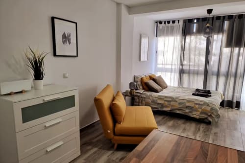 Moderno apartamento estudio en Fuengirola con silla naranja, tocador blanco y vistas a la ciudad por amplios ventanales