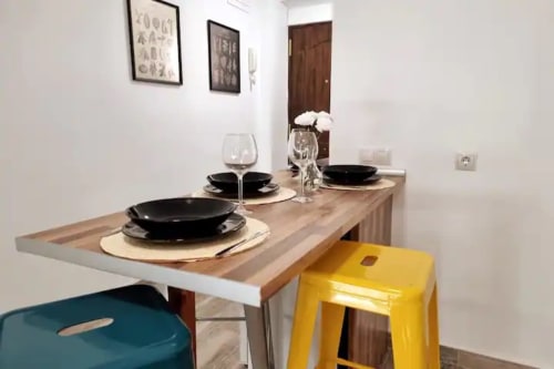 Zona de comedor de estudio moderno en Fuengirola con mesa de madera, platos negros, taburete amarillo y vistas a la ciudad