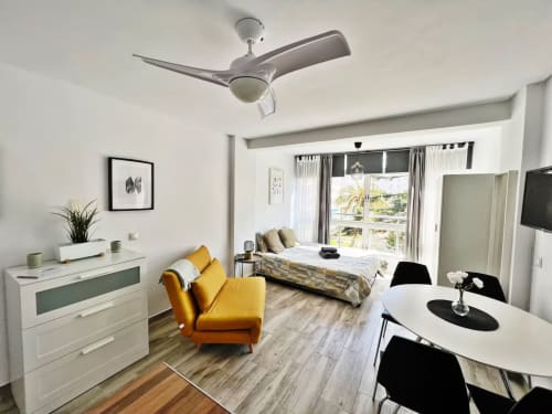 Moderno apartamento estudio en Fuengirola con sillón amarillo, cama, cómoda blanca, ventilador de techo y vistas a la ciudad