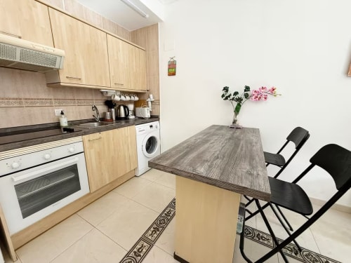 Estudio-cocina moderno en Fuengirola con muebles de madera clara, mesa de comedor, flores rosas y sillas negras