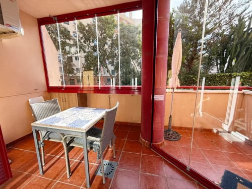 Luminosa terraza de estudio en Fuengirola con mesa de comedor, ventanas marco rojo, vistas a la ciudad y plantas tropicales