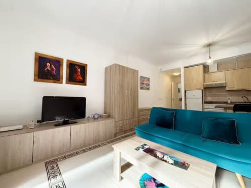 Luminoso apartaestudio en Fuengirola con sofá turquesa, televisor, cocina americana y muebles de madera