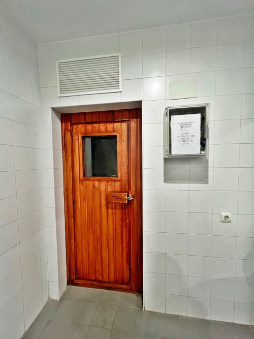 Puerta de entrada de madera con ventana y tablón de información del edificio en el pasillo blanco de azulejos del apartamento de Fuengirola