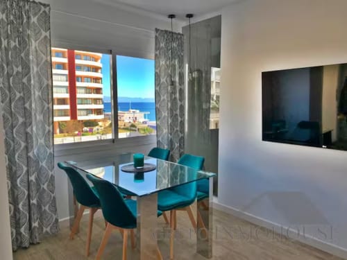 Comedor moderno en apartamento de Fuengirola con vistas al mar, mesa de cristal, sillas turquesas y televisor de pantalla plana