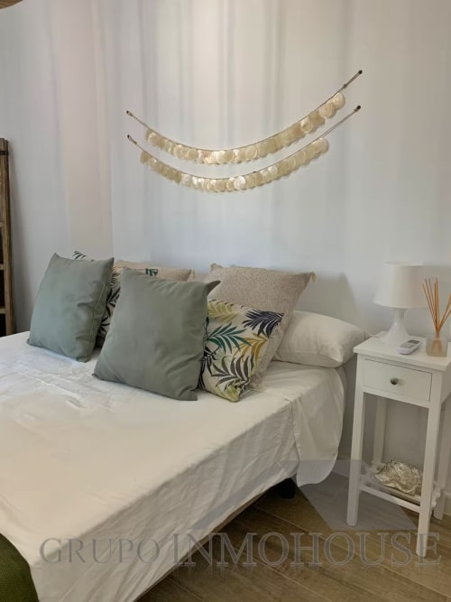 Dormitorio de apartamento frente al mar en Fuengirola con vista al mar, cama blanca, cojines decorativos y guirnalda de monedas doradas