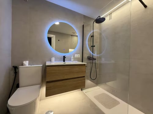 Baño moderno en apartamento frente al mar, Fuengirola. Espejo LED, tocador de madera, ducha con mampara de cristal.