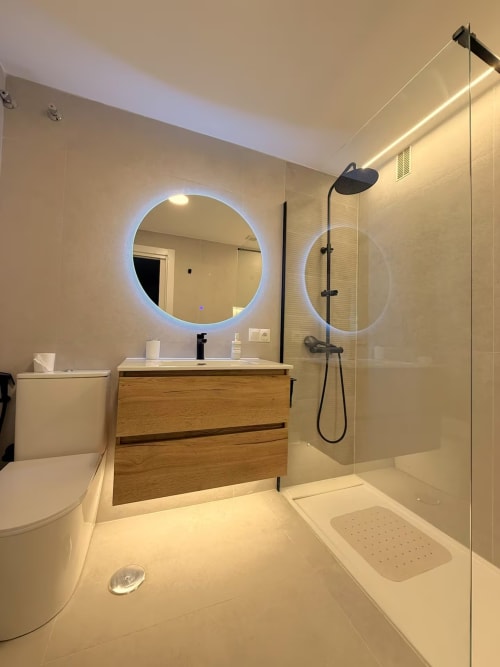 Baño moderno frente al mar en Fuengirola con espejo LED, tocador de madera y ducha abierta