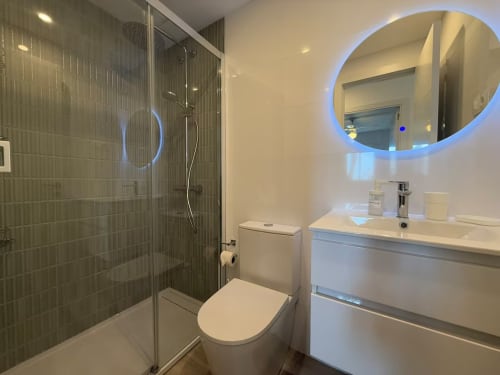 Baño moderno en apartamento Grupo Inmo House Fuengirola con ducha de cristal, espejo circular iluminado y tocador blanco.