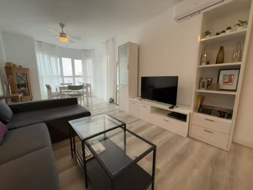 Luminoso salón del apartamento en Fuengirola con televisor, estanterías, sofá negro, mesa de cristal y vistas al balcón