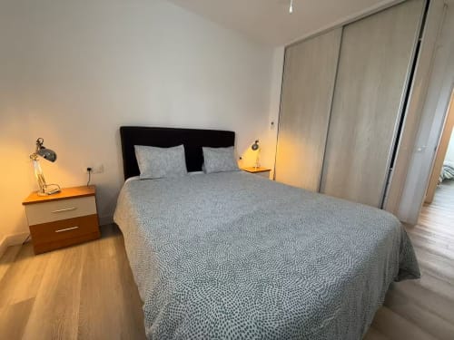 Dormitorio moderno en apartamento de playa de Fuengirola con vistas a piscina, ropa de cama gris, cabecero negro y mesillas de madera