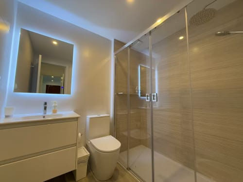 Baño moderno en apartamento de playa en Fuengirola con espejo iluminado, lavabo flotante, mampara de vidrio y ducha de lluvia