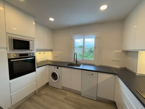 Cocina moderna con armarios blancos, encimeras oscuras, electrodomésticos de acero inoxidable y vistas al jardín en apartamento de Fuengirola