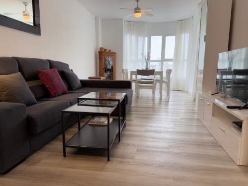 Sala moderna en apartamento de playa en Fuengirola con sofá oscuro, mesas de café, comedor y vistas al balcón