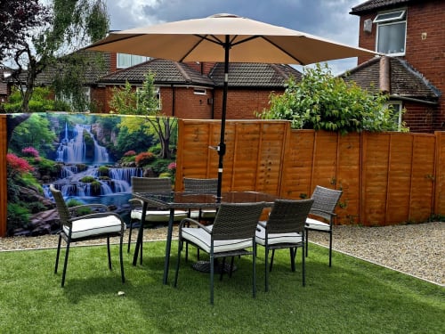 Garden seating area at Leeds Luxurious Stays villa with parasol, wicker chairs, and waterfall backdrop panel