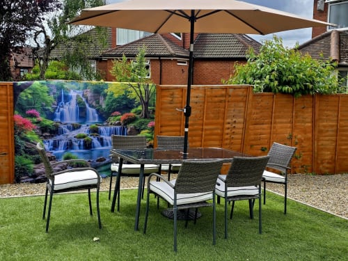 Garden patio at Leeds Luxurious Stays villa with dining table, wicker chairs, umbrella, and waterfall artwork.
