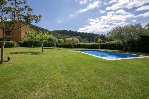 Amplio jardín con piscina rectangular, césped cuidado, olivos y casa de piedra en la villa La Pastora