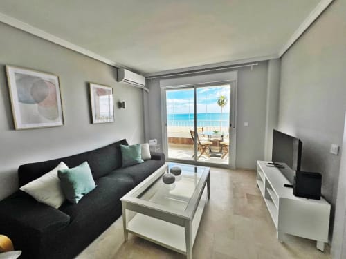 Moderno apartamento frente al mar en Fuengirola con vistas al mar, sofá oscuro, mesa de cristal, TV y balcón con vista a la costa