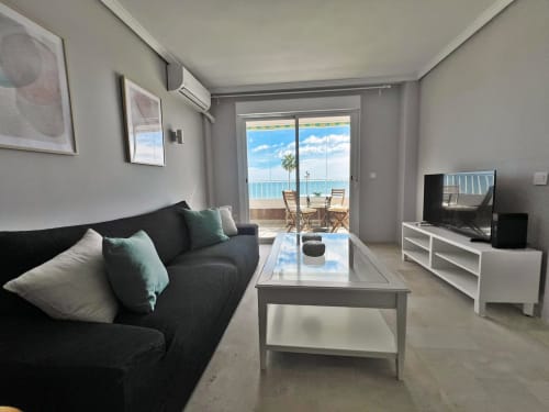 Salón de apartamento de playa moderna en Fuengirola con vistas al mar, sofá negro, TV de pantalla plana y acceso a terraza