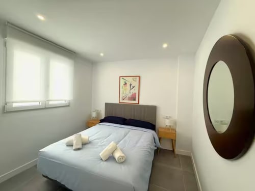 Moderno dormitorio en Fuengirola con cama queen, espejo redondo, mesillas de madera y ventanas luminosas