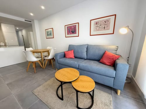 Moderno salón de apartamento en Fuengirola con sofá azul, cojines rojos, mesas de madera y cuadros enmarcados en paredes blancas