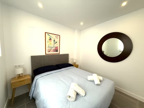Dormitorio moderno en apartamento de playa en Fuengirola con cabecero gris, mesilla de madera, espejo redondo y ropa de cama blanca