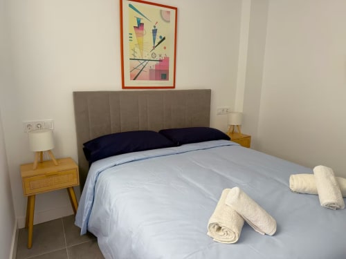 Dormitorio moderno y luminoso en apartamento de playa de Fuengirola con cabecero gris, ropa de cama blanca, almohadas oscuras y mesitas de noche de madera