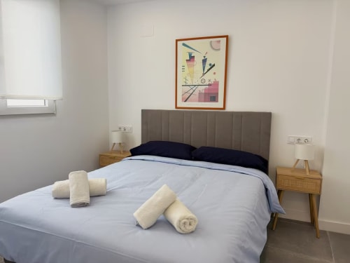 Dormitorio moderno en apartamento de playa Fuengirola con cabecero tapizado gris, mesitas de madera y toallas de bienvenida en la cama