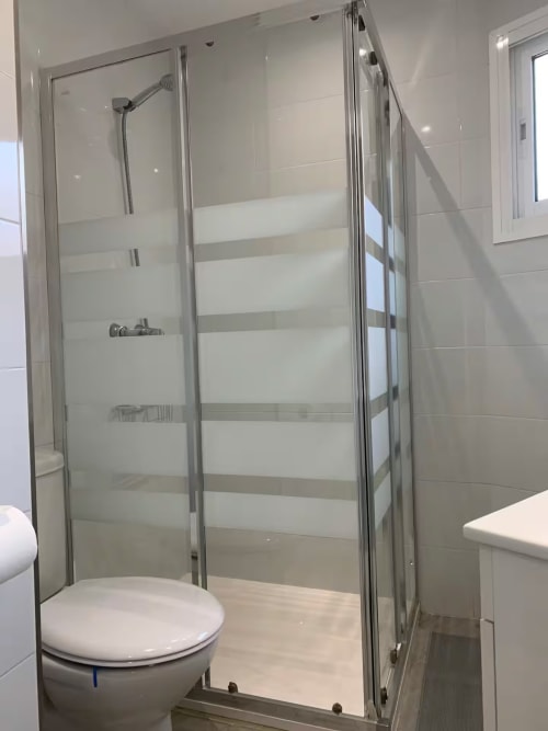 Baño moderno con ducha a ráiz, cristalera y accesorios blancos en apartamento de Fuengirola