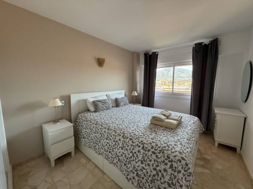 Dormitorio luminoso con vistas a las montañas en apartamento de Fuengirola, muebles blancos, ropa de cama estampada, cortinas oscuras
