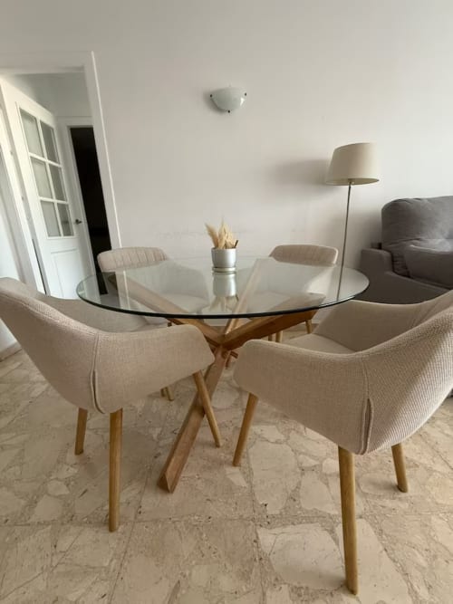 Comedor moderno con mesa redonda de cristal, sillas beige y base de madera en apartamento de playa en Fuengirola