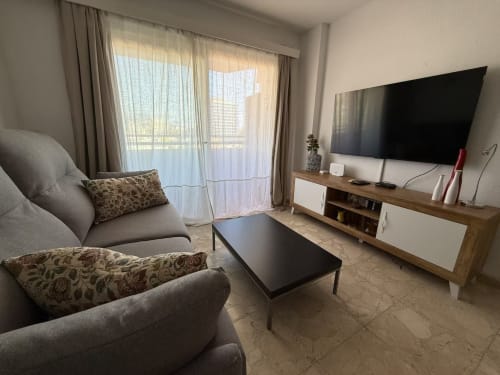 Moderno salón en Fuengirola con sofá gris, televisor de pantalla plana, mesa de café negra y balcón con vistas a montaña