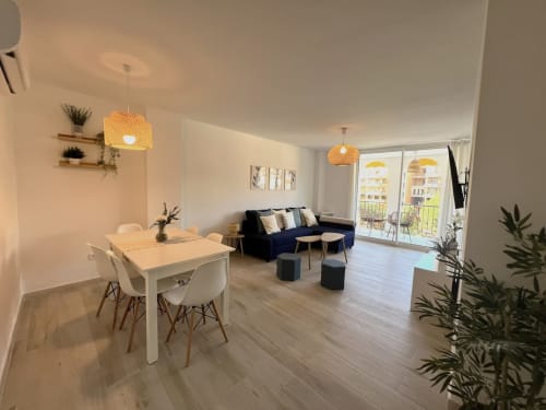 Luminoso apartamento de playa en Fuengirola con zona de comedor moderna, sofá azul marino, lámparas colgantes tejidas y balcón con vistas al jardín