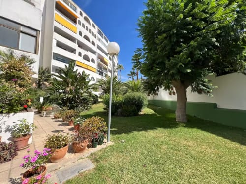 Luminoso patio ajardinado en apartamento de playa en Fuengirola con exuberante vegetación, flores en macetas y entrada moderna del edificio