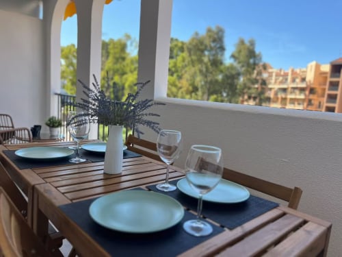 Elegante área de comedor al aire libre en balcón de apartamento frente al mar en Fuengirola con mesa preparada, vistas al jardín y blanco moderno