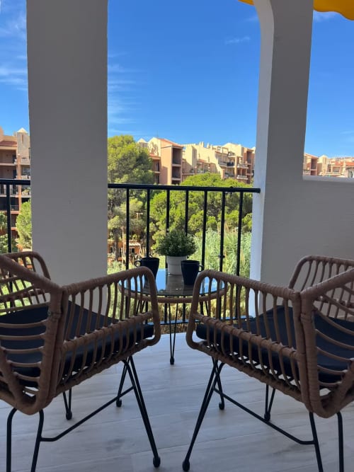 Balcón de apartamento en Fuengirola con sillas de mimbre con vista al jardín y edificios amarillos bajo cielo azul despejado