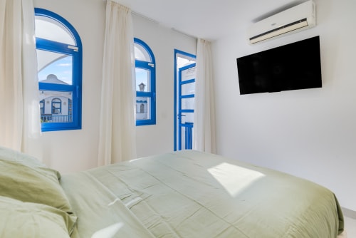 Dormitorio luminoso con ventanas arqueadas de marco azul con vistas a Maspalomas, TV de pared y ropa de cama verde pálido en FLOR DEL